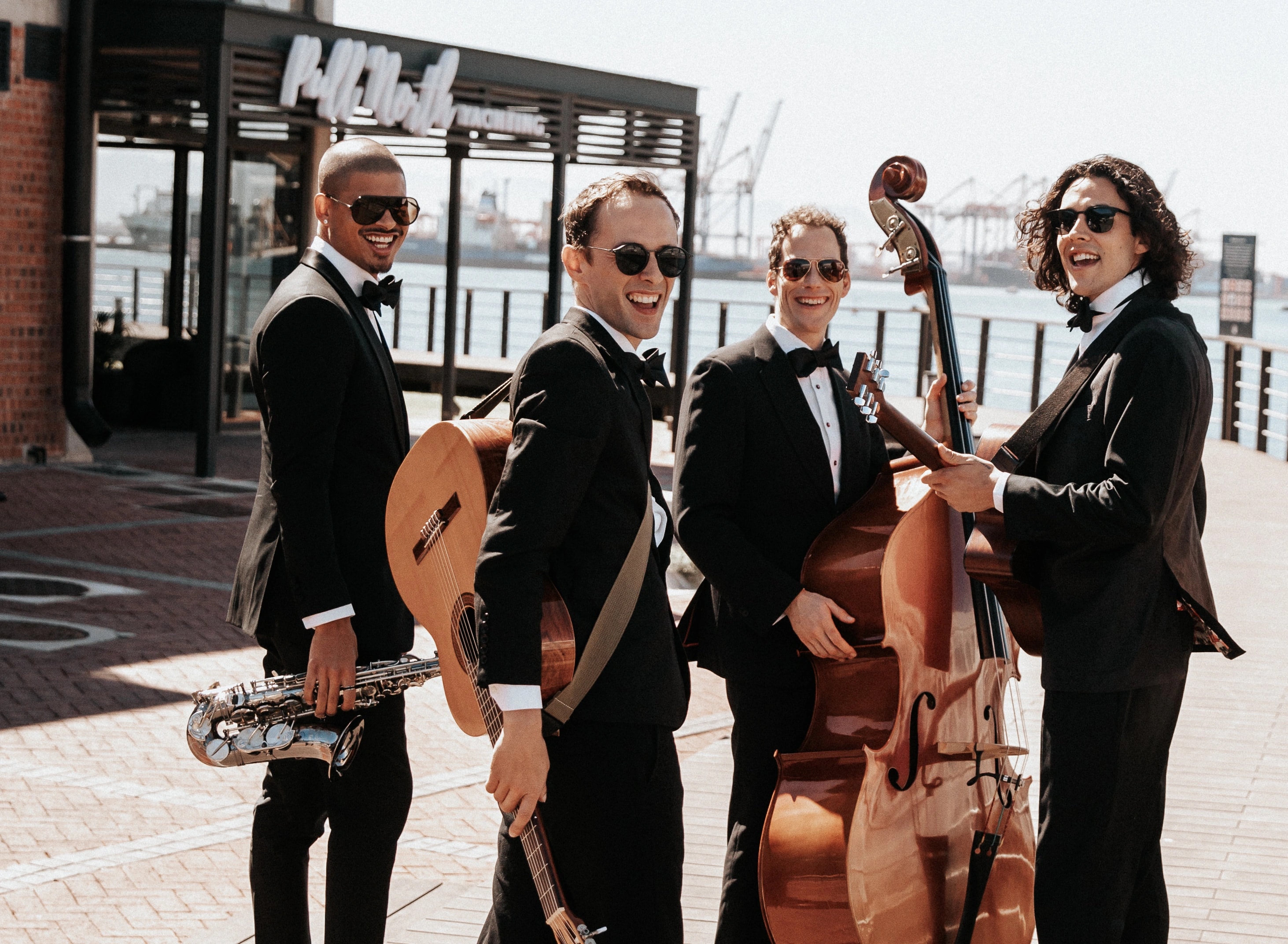 Four Empire musicians in black tie with saxophone, guitars and double bass on a waterfront boardwalk